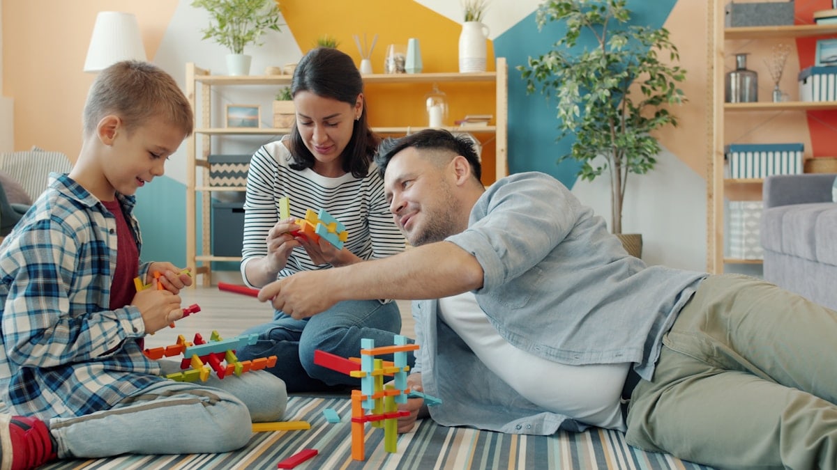 Child is playing with mother and father cheerful adults at home building with construction blocks sitting on floor together. Youth and family concept.